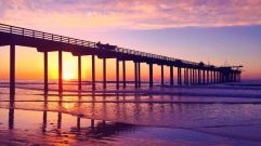 The Sun sets over the ocean in the background, and a pier is covered in shadows in the foreground.